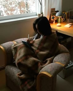 a woman wrapped up in a blanket sitting on a chair next to a desk and window