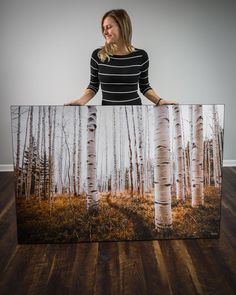 a woman is holding up a painting with white birch trees in the woods behind her