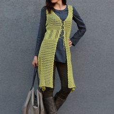 a woman standing next to a gray wall holding a purse and wearing a yellow knitted vest