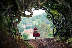 a woman in a red dress walking down a dirt road with trees on both sides