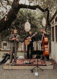 three men are playing music in front of an outdoor area with trees and string lights