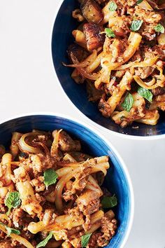 two blue bowls filled with pasta and meat on top of a white table next to silverware