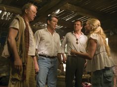 three men and two women are standing in a barn talking to each other while the man is wearing a white shirt