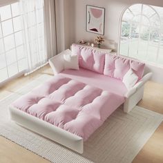 a white and pink bed with pillows on it in a living room next to a window