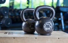 two black kettles sitting on top of a wooden table in a gym with rows of dumbbells behind them