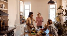 three women standing around a kitchen table with food on it and one woman holding a cup