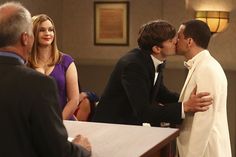 two men in suits kissing each other at a table with people sitting around and looking on