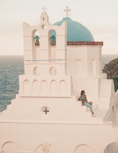 a man sitting on the side of a white building next to the ocean with a blue dome