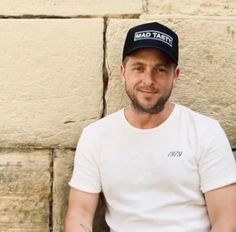 a man wearing a hat sitting in front of a stone wall with writing on it