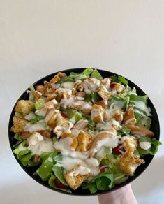 a hand holding a black bowl filled with salad and dressing on top of white table