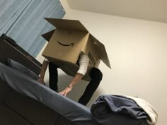 a person standing on top of a bed with a cardboard box on their head and an amazon box on his head