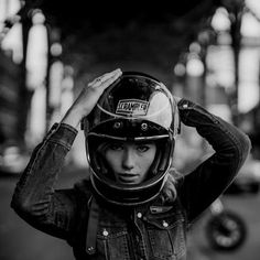 a woman wearing a motorcycle helmet and denim jacket with her hands on her head while standing in the street