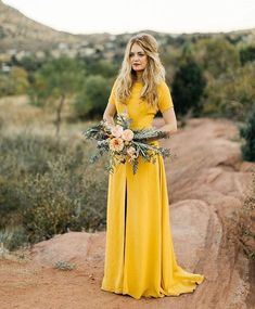 a woman in a yellow dress standing on top of a dirt road with flowers and greenery