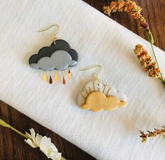 two earrings with clouds hanging from them on a table next to dried flowers and paper towels