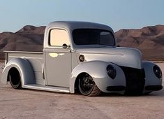 an old silver truck parked on top of a dirt field in front of some mountains