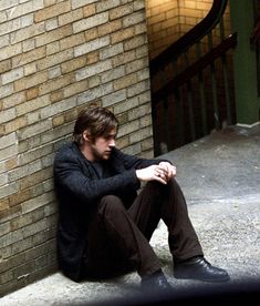 a man sitting on the ground next to a brick wall with his hands in his pockets