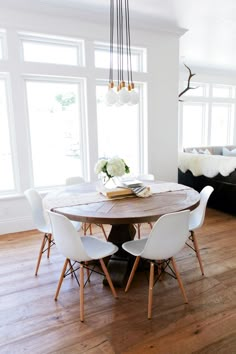 a dining room table with white chairs in front of large windows and wooden flooring