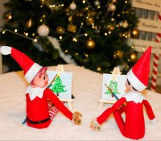 two elfs are sitting in front of a christmas tree, one is holding a paintbrush