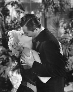 a man and woman are kissing in front of some plants, one is holding a teddy bear