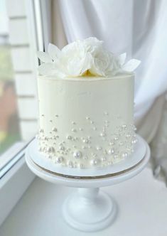 a white cake with flowers and pearls on a table next to a window sill