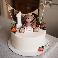 a birthday cake with a teddy bear on top and berries around the base, along with a number one sign