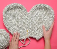 someone crocheting a heart on top of a pink surface next to two balls of yarn