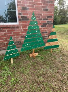 three wooden christmas trees sitting in front of a brick building with grass on the ground