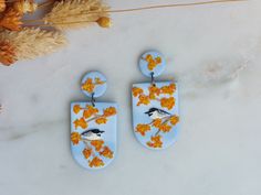 two earrings with birds on them sitting on a marble table next to dried flowers and leaves