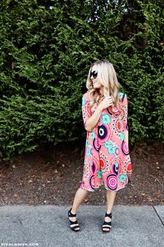 a woman standing in front of some bushes wearing sunglasses and a pink dress with flowers on it