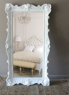 a white bed sitting under a mirror next to a lamp and a wall with a chandelier