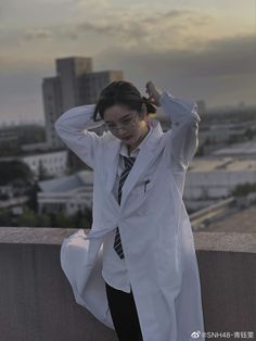 a woman wearing a white coat and tie standing on top of a building with her hands behind her head