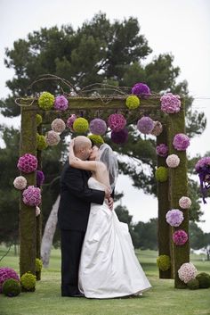 a bride and groom kissing under an archway decorated with pink and purple flowers at their wedding