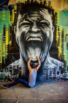 a man sitting on the ground in front of a wall with a painting of a screaming face