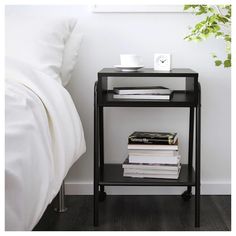 a black end table with some books on it next to a white bed and plant