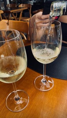 two wine glasses sitting on top of a wooden table next to each other with white wine being poured into them