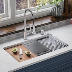 a kitchen sink under a window next to a cutting board and knife holder with fruit on it