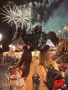 two girls are looking at fireworks in the sky over an amusement park with mickey ears on their heads