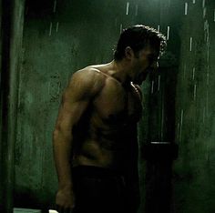 a shirtless man standing next to a toilet in a dark room with rain falling down on the walls