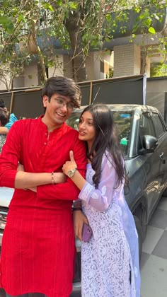 two people standing next to each other in front of a parked car and looking at the camera