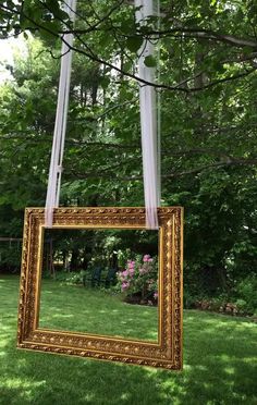 a gold framed mirror hanging from a tree in the middle of a yard with pink flowers