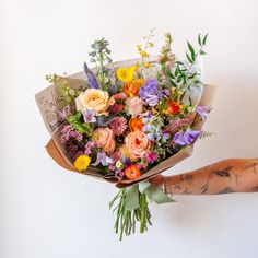 A tattooed arm holds a bouquet wrapped in floral newspaper and brown kraft paper in front of a white backdrop. It's filled with pink, orange, yellow, and purple flowers, including piccolini daisies, roses, veronica, sweet pea, clematis, delphinium, oncidium orchid, and lisianthus. Wildflower Bridal Bouquets, Wildflower Wedding Bouquet, Summer Wedding Bouquets, Boquette Flowers, Spring Wedding Flowers, Summer Bouquet, Colorful Bouquet, Wildflower Bouquet, Beautiful Bouquet Of Flowers