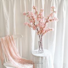 a vase with pink flowers on a table next to a white chair and drapes