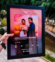 a person holding up a framed photo in front of trees and grass, with the image of a bride and groom on it