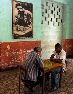 two men sitting at a table playing chess in front of a painting on the wall