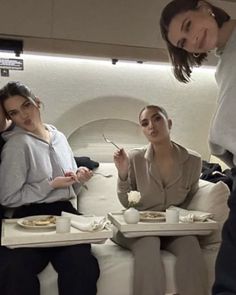 three women sitting on a couch eating food from trays in the middle of an airplane