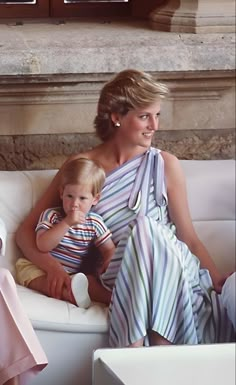 a woman sitting on top of a white couch next to a little boy in a sling