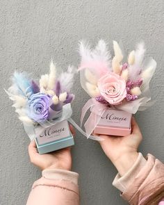 two small boxes with flowers and feathers are held by someone's hands in front of a gray wall