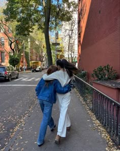 two women are walking down the sidewalk in front of a red brick building and trees