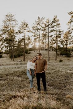 a man and woman standing in the middle of a field with pine trees behind them