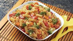 a bowl of pasta with broccoli and tomatoes in it on a mat next to yellow utensils